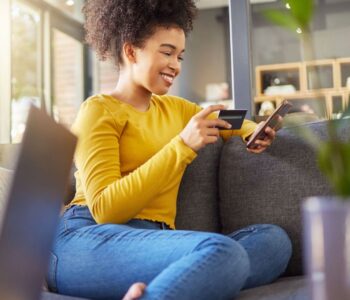 A Woman wearing a yellow jersey and a blue jeans using a credit card wisely with a phone on a counch