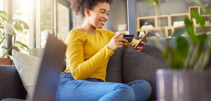 A Woman wearing a yellow jersey and a blue jeans using a credit card wisely with a phone on a counch